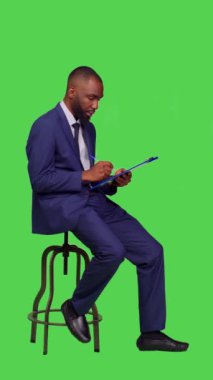 Dikey video: Afro-Amerikan bir adamın kağıtlar üzerinde iş notları düşünmesi, pano dosyaları üzerinde yeni fikirler üretmesi. Stüdyoda sandalyede oturan genç iş adamı,