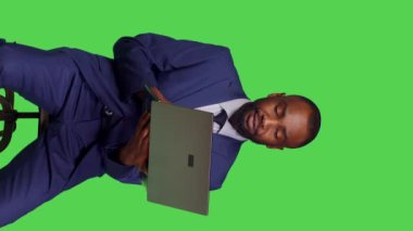 Dikey video: Afrikalı Amerikalı yetişkinlerin koltukta oturup dizüstü bilgisayarla internet sitesi üzerinde çalışırken bilgisayar kullanışının ön görüntüsü. Resmi takım elbiseli şirket yöneticisi yeşil ekran üzerinde kablosuz bilgisayar tutuyor