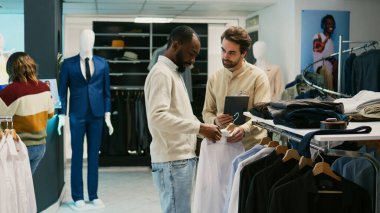 Afro-amerikalı adam askıları kontrol ediyor, mağaza asistanına yeni moda koleksiyonunu soruyor. Alışveriş merkezi çalışanıyla perakende satış mağazasındaki giysiler hakkında konuşan genç bir müşteri..