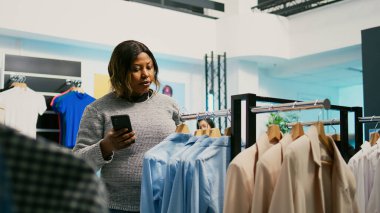 Mağazadaki kadın müşteri web sitesi stoklarını kontrol ediyor, modern online ürünlere bakmak için cep telefonu kullanıyor. Alışverişkolik genç yetişkin yeni moda eğilimlerini, ticari faaliyetleri inceliyor.