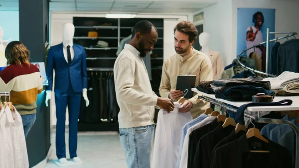 ハンガーに商品をチェックするアフリカ系アメリカ人男性 新しいファッションコレクションについて店員に尋ねる 小売店のブティックで服についてモールの従業員と話す若いクライアント — ストック写真