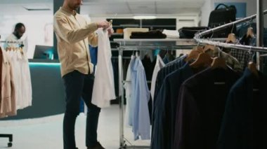 Erkek müşteri butikte moda koleksiyonunu ve resmi kıyafetleri inceliyor. Genç yetişkin kıyafetleri için kıyafet alışverişi yapıyor. Mağazada asılı duran mallara bakan beyaz bir adam..