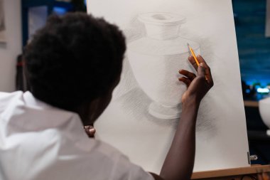 Afro-Amerikan bir adamın resim stüdyosundaki tuvale kurşun kalemle çizdiği resimlere yakından bakın. Erkek genç sanat öğrencisi sanatsal atölyede boş tuval üzerinde vazo çiziyor..