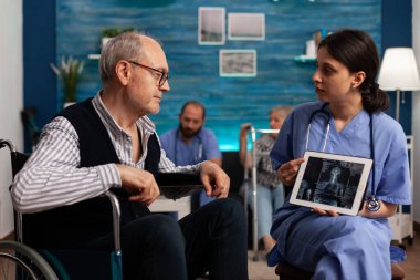 Fizik tedavi ve rehabilitasyon uzmanı tekerlekli sandalyedeki yaşlı hastaya vücut taraması görüntüleriyle ekranı gösteriyor. Özel merkezdeki tıbbi randevuda engelli bir adam..