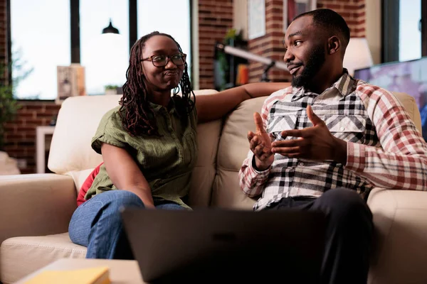 アフリカ系アメリカ人の夫婦は 自宅のリビングルームでソファに座っている間 お互いに話をします ガールフレンド見て不満を説明し 手ジェスチャーでテーブルの前でラップトップ — ストック写真