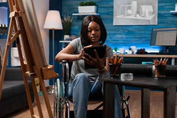 African american female art student wheelchair user taking remote art classes with tablet in home crafts studio. Young drawing schoolgirl using digital tablet to learn new sketching techniques.