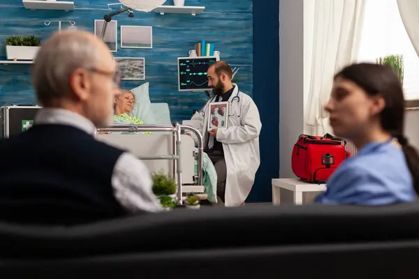 医生用数码平板电脑向在医院病床上的老年女性病人解释心脏功能 丈夫坐在沙发上与医疗助理谈论治疗问题 — 图库照片