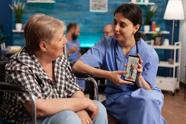 Female nurse in scrubs holding phone for online medical consultation of senior female patient in wheelchair. Offering assistance with technology management in a nursing home.