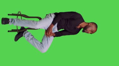 Dikey video: Sandalyedeki Afro-Amerikan adam özgüvenle poz veriyor, tüm vücudu yeşil ekranda oturuyor. Genç erkek model kendini iyimser hissediyor ve stüdyoda bir şeyler bekliyor.