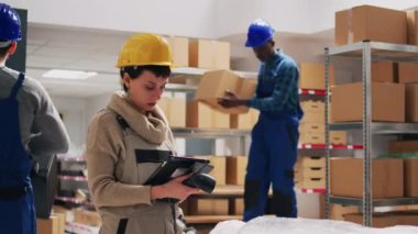 Kadın denetçi, depo deposunda tarayıcı ve tablet kullanarak envanter için kutularda ürünleri tarıyor. Genç yetişkin işçilerle lojistik ve planlama siparişi nakliyatı üzerinde çalışıyor..
