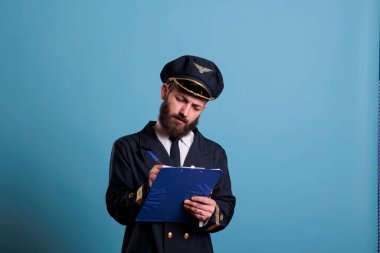 Uçak pilotu pano tutuyor, yazıyor, belgeli bir havacı. Profesyonel üniformalı bir uçak kaptanı. Havalimanı önünde form dolduruyor. Mavi arka planda orta ölçekli stüdyo çekimi.