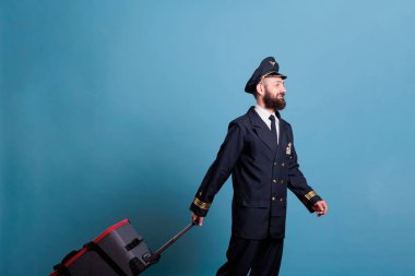Profesyonel havayolu üniformalı bir uçak kaptanı bavulla yürüyor. Bavullu gülümseyen pilot uçuşa hazır, havaalanında yürüyor, havacı bagajını tutuyor, yandan bakıyor.