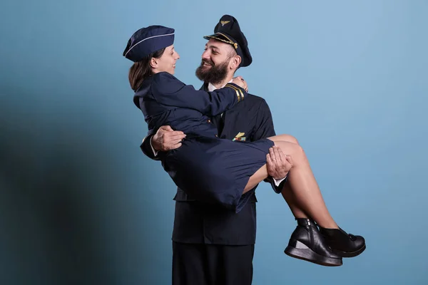 Pilot holding stewardess in arms, looking at each other portrait, aircrew in professional uniform. Happy aviator and flight attendant romantic couple in love studio medium shot