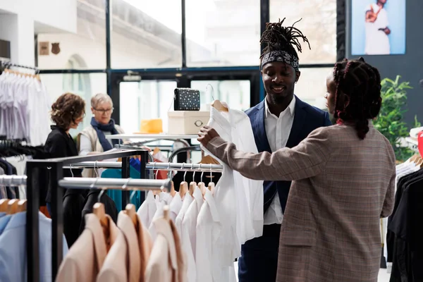 ショールームの従業員は 商業活動中に白いシャツを選択するアフリカ系アメリカ人のクライアントを支援しています ショッピングセンターでファッショナブルな服を購入エレガントな顧客 ファッションコンセプト — ストック写真