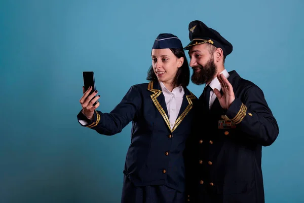 Smiling copilot and air hostess talking on smartphone online videocall together. Airlane captain and flight attendant chatting on mobile phone videoconference, answering call, studio shot