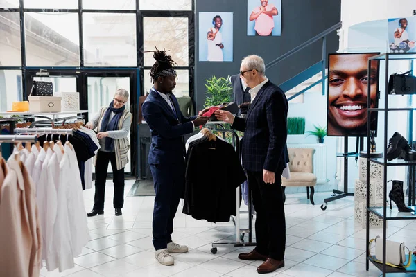Stylish worker helping senior client with red tie, analyzing fabric in modern boutique. Elderly client wearing formal suit shopping for casual wear and fashionable merchandise in showroom