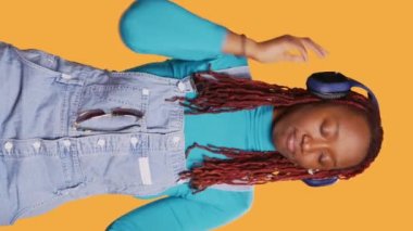 Dikey video: mp3 müzik eşliğinde dans eden neşeli kız, stüdyoda şarkı dinlemek için kulaklık kullanıyor. Genç, gülümseyen, dans eden, eğlenen, tatilde sesler dinleyen biri. El bilgisayarı