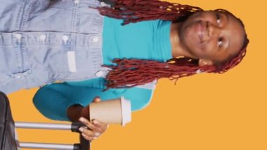 Dikey video: Kamerada gülümseyen gezgin bir fincan kahve içiyor, bavullar ve bavullarla seyahate çıkmaya hazırlanıyor. Heyecanlı insan tatil yerine gitmeden önce içkinin tadını çıkarıyor.