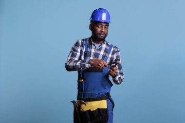 Afro-Amerikalı inşaatçı mavi arka planda kerpeten kullanırken konsantre oluyor. İş aletleri, koruyucu kask ve tulum kullanan işçilerin stüdyo portresi.