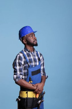 Afrika kökenli Amerikalı müteahhit dikkatle yukarı bakıyor, işi yapmak için gerekli araç ve gereçleri detaylandırıyor. Stüdyoda mavi arka planda çekilen iş tulumu ve şapka giyen inşaat işçisi..