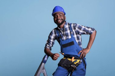 İyimser müteahhit, mavi arka plandaki işi bitirdikten sonra dinlenirken kameraya büyük bir gülümsemeyle bakıyor. İnşaat işleri için gerekli alet kemerini kullanan müteahhit.