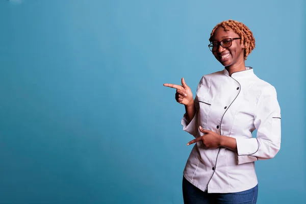 Horizontal shot of happy african american woman enjoying herself in studio shot on blue background pointing to free space for advertising content. Cook in uniform looking optimistic.