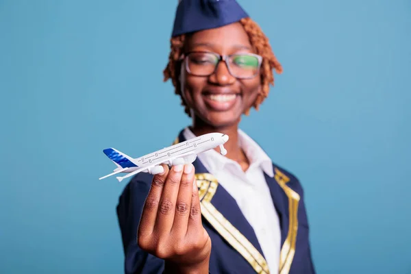 Optimistic african american flight attendant in uniform playing with small custom made airplane toy. Female crew member having some fun before starting a long flight around the world.