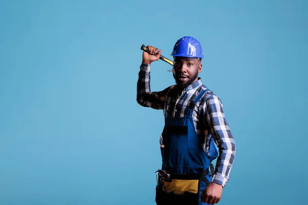 African american construction worker aggressively holding hammer looking at camera. Builder in construction helmet and overalls, looks angry isolated on blue background.