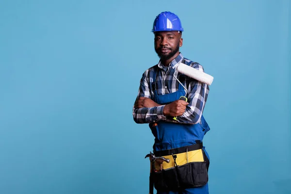 Professional painter looks at camera holding paint roller with overalls and protective helmet, arms crossed over chest. African American construction worker against blue background in studio shot.