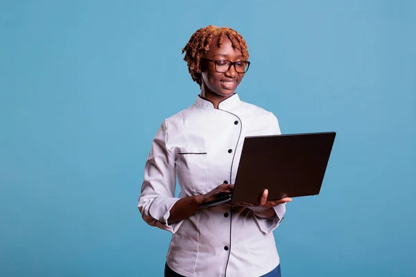 アフリカ系アメリカ人の女性シェフは コンピュータを使用して インターネット上でレシピを検索 閲覧します 女性のアフロヘア調理器のスタジオ肖像 ビデオ通話を動作させるためにラップトップを使用して — ストック写真