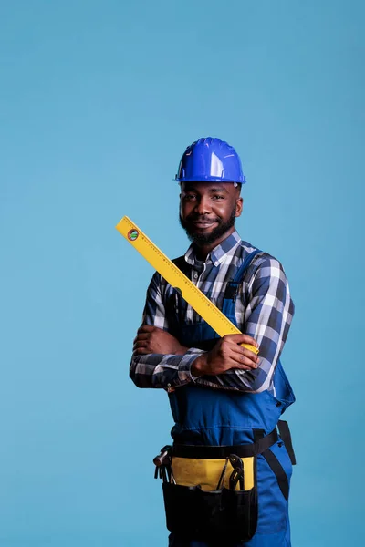 Smiling african american repair worker in uniform holding tape measure on blue studio background. Experienced builder holding work tools wearing coveralls and hard hat.