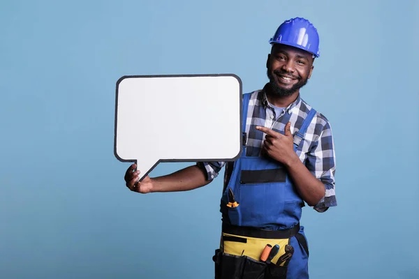 Optimistic african american man pointing to empty white dialogue bubble with copy space, showing advertising mockup. Cheerful construction worker holding speech frame, against blue background.