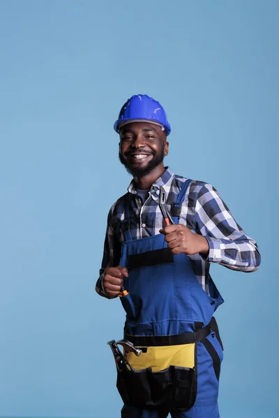 Excited african american man in hard hat showing thumbs up as sign of approval. Optimistic builder happy at work wearing belt with tools and uniform, construction and renovations.