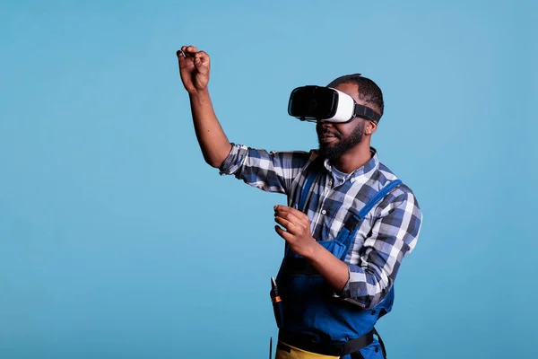 Expert in renovations looking at 3d plans with virtual reality goggles. Contractor using modern technology to apply it in construction business, dressed in uniform against blue background.