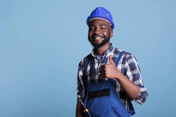 誇り高いアフリカ系アメリカ人のプロの電気技師は カバーと保護ヘルメットを着用 青を背景に笑顔で作品に興奮を感じながら親指を表示します — ストック写真