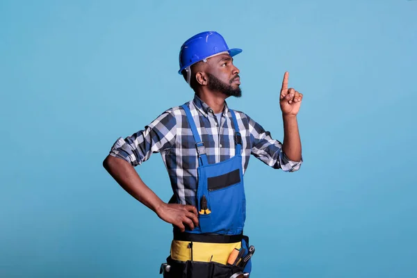 アフリカ系アメリカ人の建築家がスタジオの上方を指しているのは 青い背景を背景に撮影し 内部の改装を行う際に作業服と硬い帽子を着用した 改修と改良の概念 — ストック写真