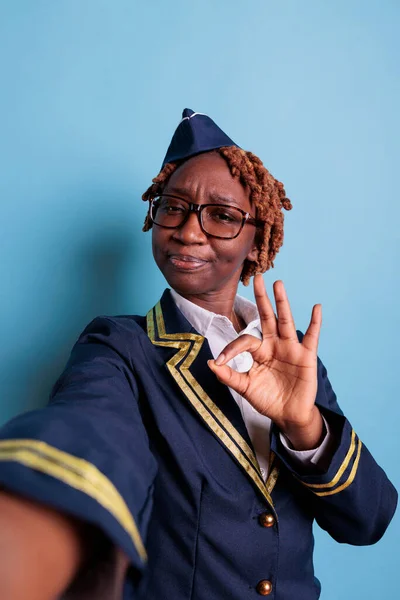 Female flight attendant showing finger gesture of approval in front of camera, feeling excited about achievement at work. Happy woman wearing uniform and glasses in studio shot.