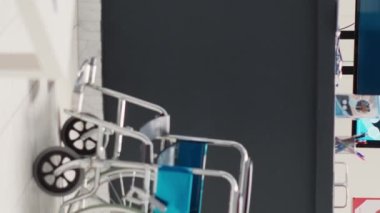 Dikey video: Modern tıp merkezinde tekerlekli sandalyesi olan hastane bekleme alanı, koltuklar ve sigorta broşürleriyle dolu boş resepsiyon masası. Tezgahı olan klinik lobisi, fiziksel engelli.