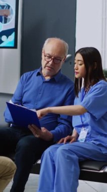 Dikey video: Hemşire yaşlı hastalara sağlık hizmetleri tedavisini hastane bekleme odasında muayene sırasında açıklıyor. Sağlık sigortası raporunu dolduran kıdemli bir adam.