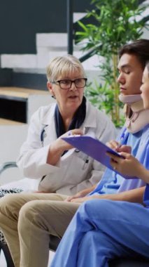 Dikey video: Sağlık görevlileri bekleme alanında muayene sırasında boyunluğu çıkarmak için yaralı bir hastayla tartışıyor. Asyalı asistan elinde pano tutuyor. Tıbbi uzmanlık yazıyor, sağlık hizmetleri planlıyor.