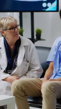 Dikey video: Yaşlı doktor hastane bekleme odasında asistanıyla tıbbi uzmanlığı açıklarken, yaralı bir hastayla sağlık tedavisini tartışıyor. Asyalı adam boyun tasması takıyor.
