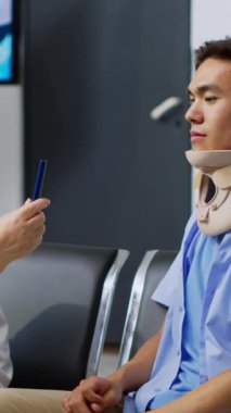 Dikey video: Yaralı hastayı muayene eden yaşlı doktor, hastane bekleme alanındaki muayene sırasında göz vizyonunu kontrol ediyor. Rahim boynu tasması takan Asyalı bir adam ilaç alıyormuş.