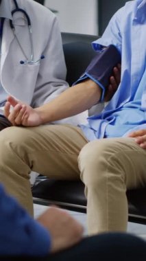 Dikey video: Hastanenin bekleme odasındaki hastanın nabız basıncını ve hipertansiyonunu ölçmek için tonometre cihazı kullanan kardiyolog. Afrikalı Amerikalı doktor Asyalı adamla kardiyoloji sınavı yapıyor.