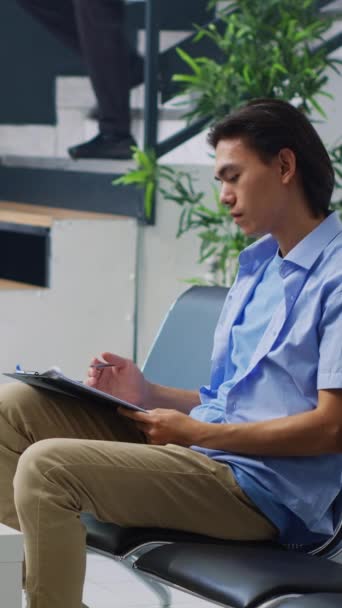 垂直ビデオ アジアの患者は 診察を受ける前に看護師に医療文書を記入し 病院の待合室で椅子に座っています 健康診断の際に保険に加入する若年成人 — ストック動画