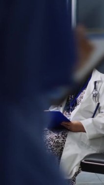 Dikey video: Hastanenin bekleme alanındaki sağlık kontrolleri sırasında hastanın tıbbi dosyaları doldurmasına yardımcı olan doktor. Adam sigorta formunu imzalarken doktor hastalık teşhisini açıklıyor.