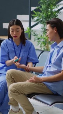 Dikey video: Ensülin seviyesini ölçen asistan ve glukometre cihazı kullanarak kan örneğinde glikoz, diyabetli Asyalı hastaya danışmanlık. Hemşire diyabetik adamla tıbbi testler yapıyor.