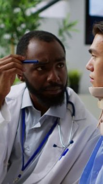 Dikey video: Hastane bekleme odasındaki muayene sırasında hastanın göz vizyonunu kontrol eden doktor, sağlık hizmetleri tedavisini açıklıyor. Asyalı adam boyun üstüne boyun tasması takıyor.