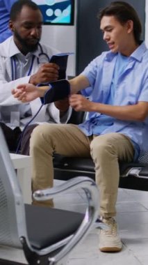 Dikey video: Hastanın nabız basıncı ve hipertansiyonunu ölçmek için tıbbi tonometre kullanan kardiyolog, hastane resepsiyonunda kardiyoloji ve kalp testi yapıyor. Sağlık görevlisi ve genç yetişkin katılımcı