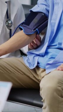 Dikey video: Kıdemli doktor Asyalı hastayla kardiyoloji muayenesi yapıyor, kan ve nabız basıncını ölçüyor. Doktor, hastanede tıbbi tonometre kullanımını kullanarak adamın hipertansiyonunu ölçüyor.