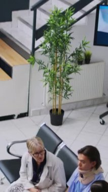 Dikey video: Hastanenin lobisine sağlık kontrolü sırasında Asyalı hemşire panoya tıbbi uzmanlık yazarken, yaşlı doktor boynu yaralanan hastayı muayene ediyor. Servikal tasma takan genç bir adam